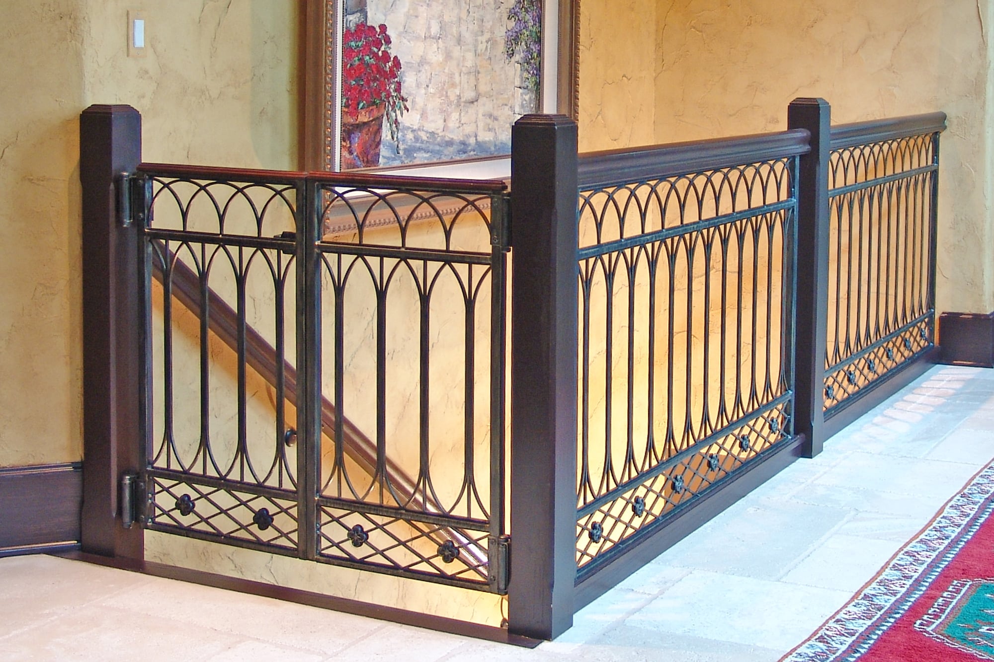 Decorative black custom ironworks safety gate and railing enclose the top of a staircase, featuring an arched pattern and geometric details, with tan walls and part of a red rug visible.