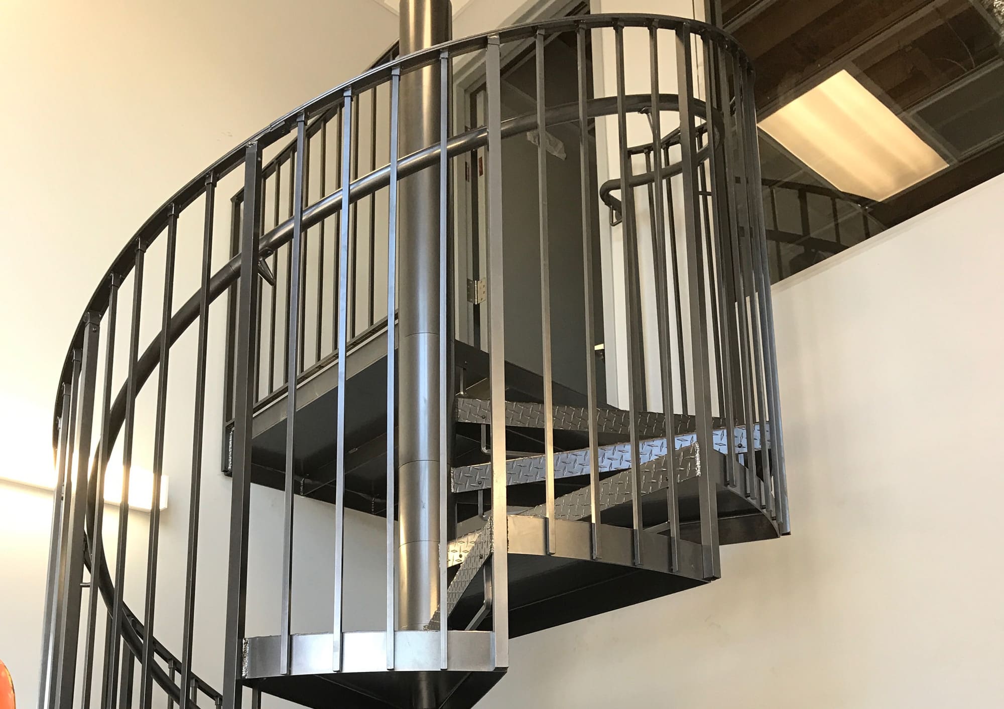 A metal spiral staircase with custom ironworks and black railings leads up to a closed door on an upper level inside a building with white walls and exposed ceiling structures.