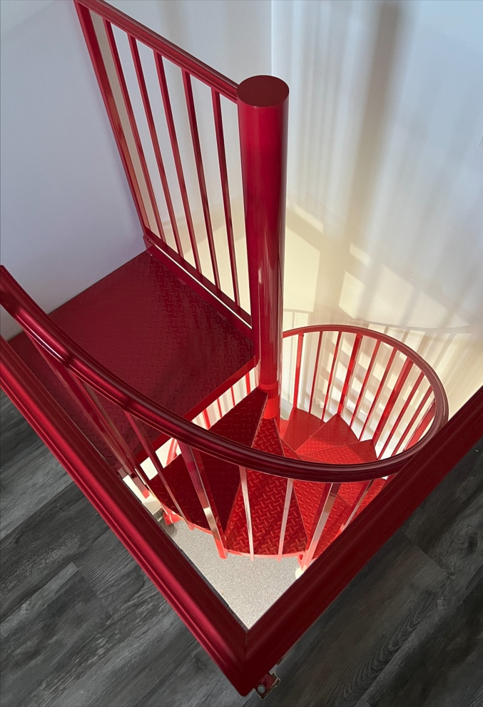 A bright red spiral staircase with custom railing and metal railings stands out against white walls and dark wood flooring, viewed from above.