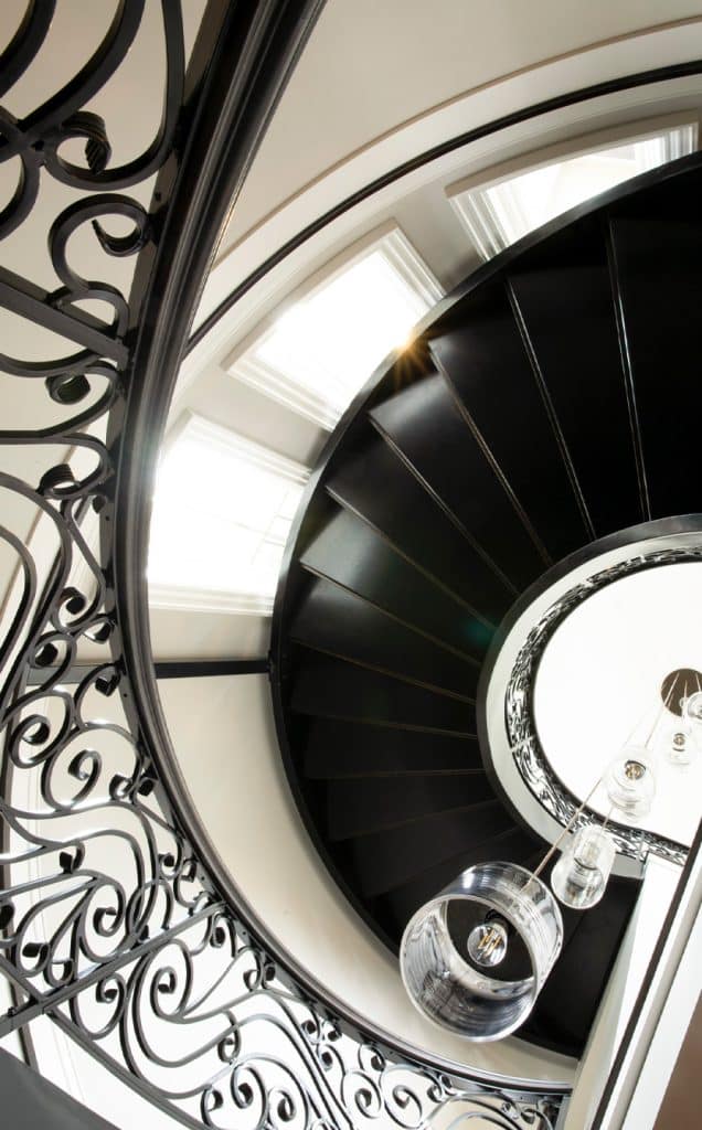 A photo looking up a black spiral staircase with custom ironworks and ornate wrought iron railings, illuminated by natural light from large windows above and modern pendant lights hanging in the center.