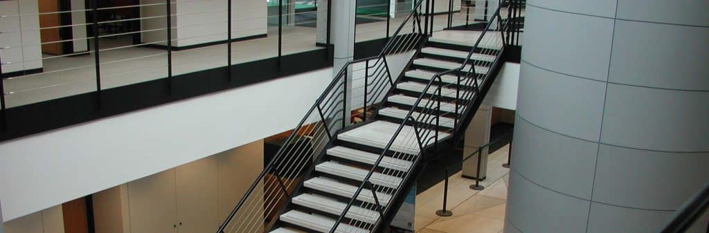 A modern indoor staircase with custom railing and white steps leads from the ground floor to an upper level in an office building with glass walls and a large cylindrical column.