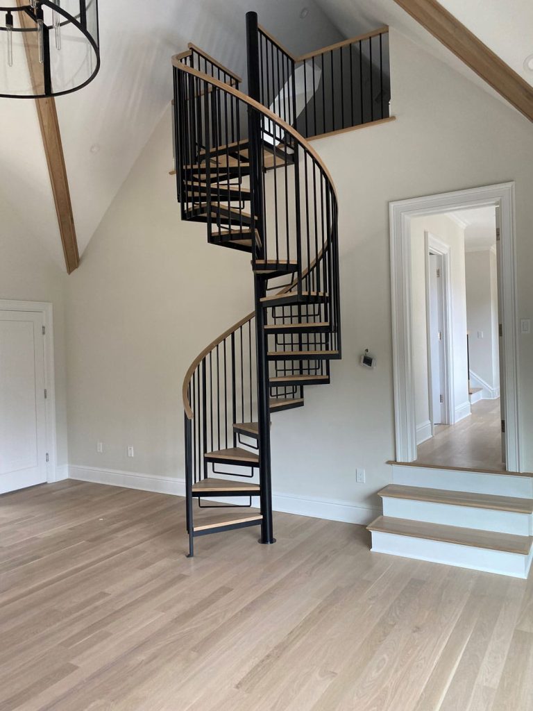 A modern room with light wood floors, white walls, and custom ironworks featuring a black spiral staircase leading to an upper loft. Wooden beams accent the sloped ceiling, and sunlight brightens the space.